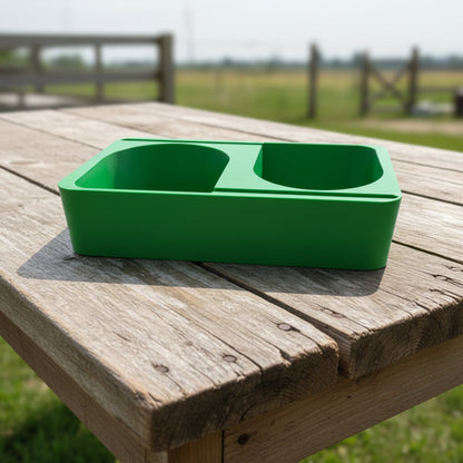 Green double-compartment feeder on a wooden table with a rural background